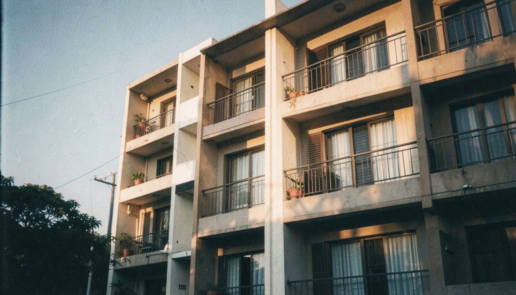 A warm analog film photograph of a modern residential apartment building facade in India, with soft golden-hour sunlight casting long shadows across balconies and windows, visible film grain, muted earth tones and pale sky